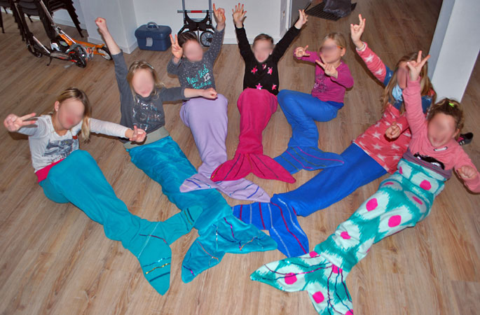 Eine Gruppe von Kindern sitzt mit selbstgenähten Kuscheldecken in Form einer Meerjungfrauenflosse im Kreis auf dem Boden und freut sich.