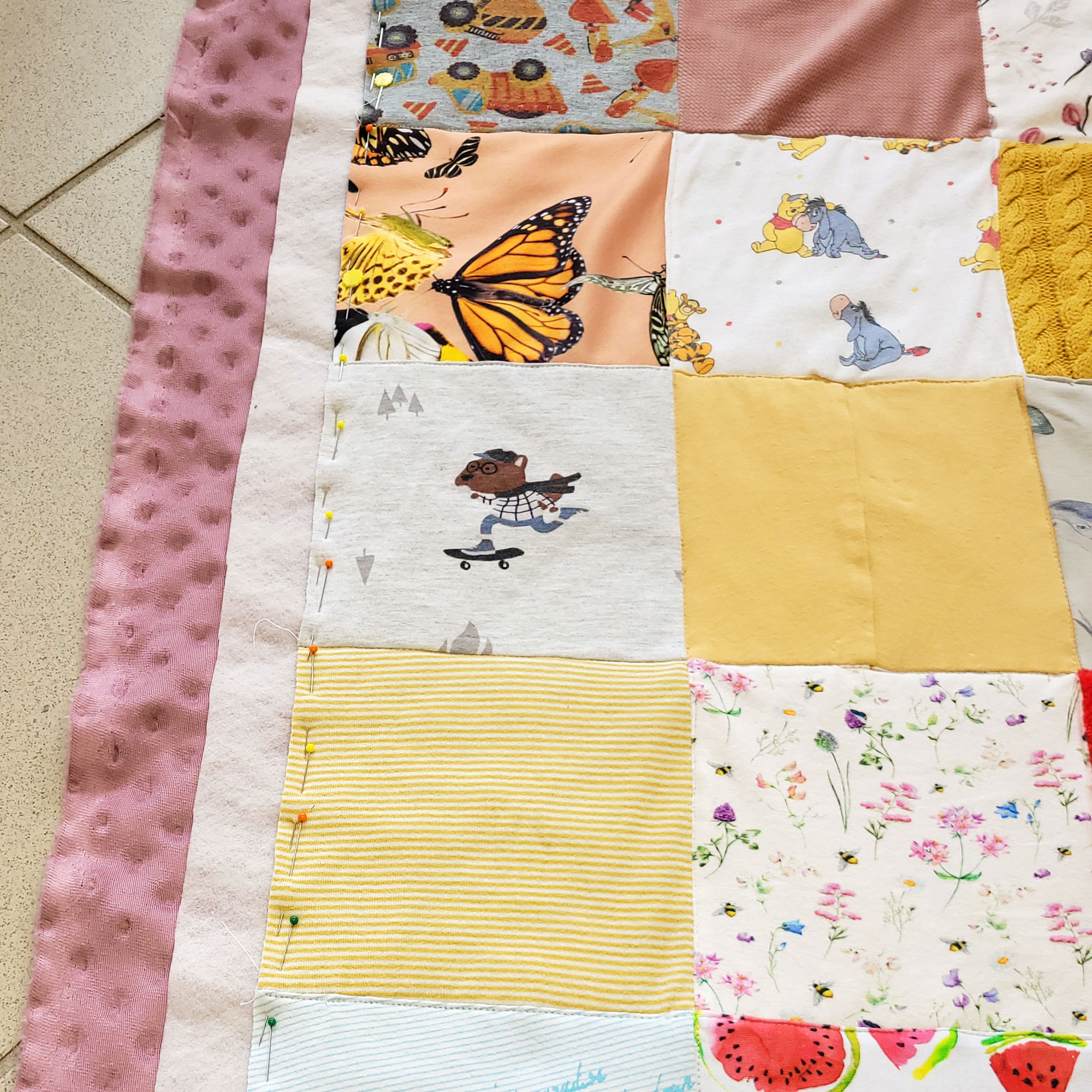 Die fertig gequiltete Decke ist vorbereitet, damit ich die Einfassung stecken kann. Die Quadrate sind am Rand gleichmäßig auf der Unterlage festgesteckt. links davon schaut ca. 4 cm breit die Vlieseline hervor. Darunter und links davon schaut weitere ca. 5 cm breit der Rückseiten Stoff hervor, bereit umgeschlagen und festgesteckt zu werden.