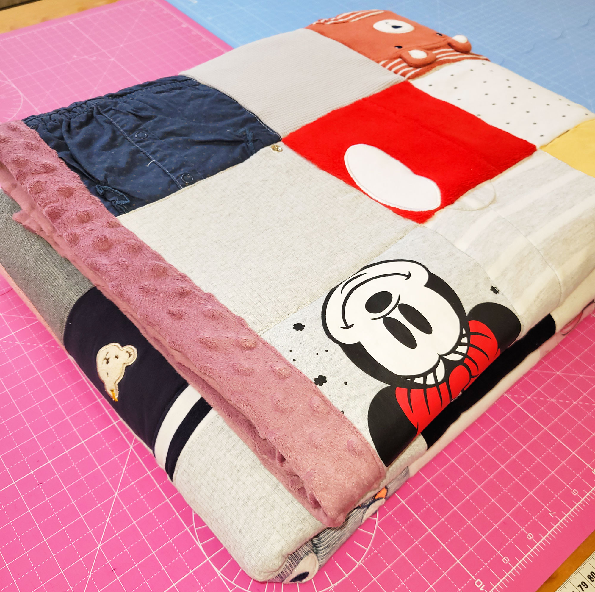 Die komplett fertige Decke liegt zusammengefaltet auf den Schneidematten meines Zuschneidetisches. Man sieht auf einem Quadrat eine strahlende Minnie Maus, ganz oben einen braun-roten Teddybären. Ein Teil der Einfassung sieht man links am Rand der Decke