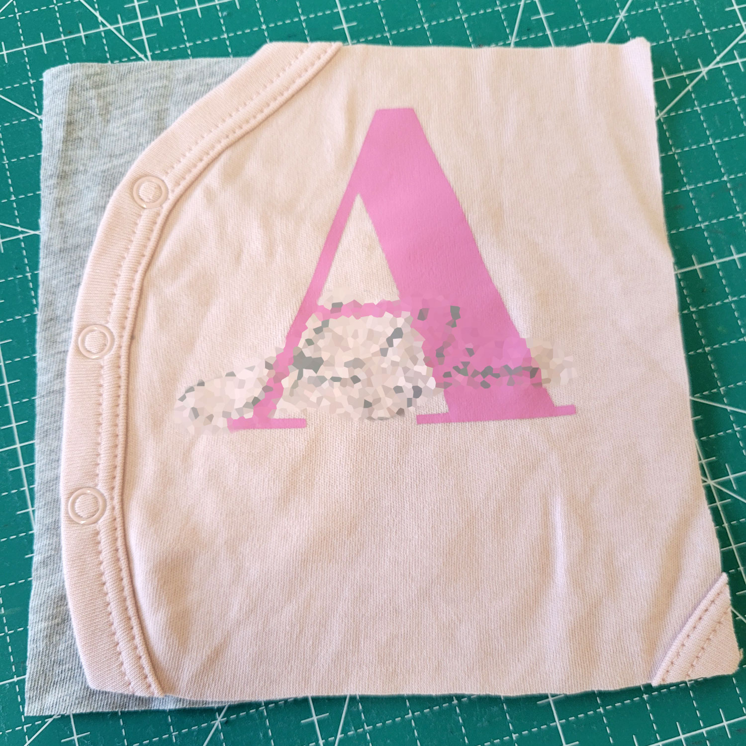 Ein ganz kleiner, rosa Babybody ist auf einem neutralen, grauen Quadrat aufgenäht. Auf dem Body befindet sich der ausgepixelte Name eines der Kinder.