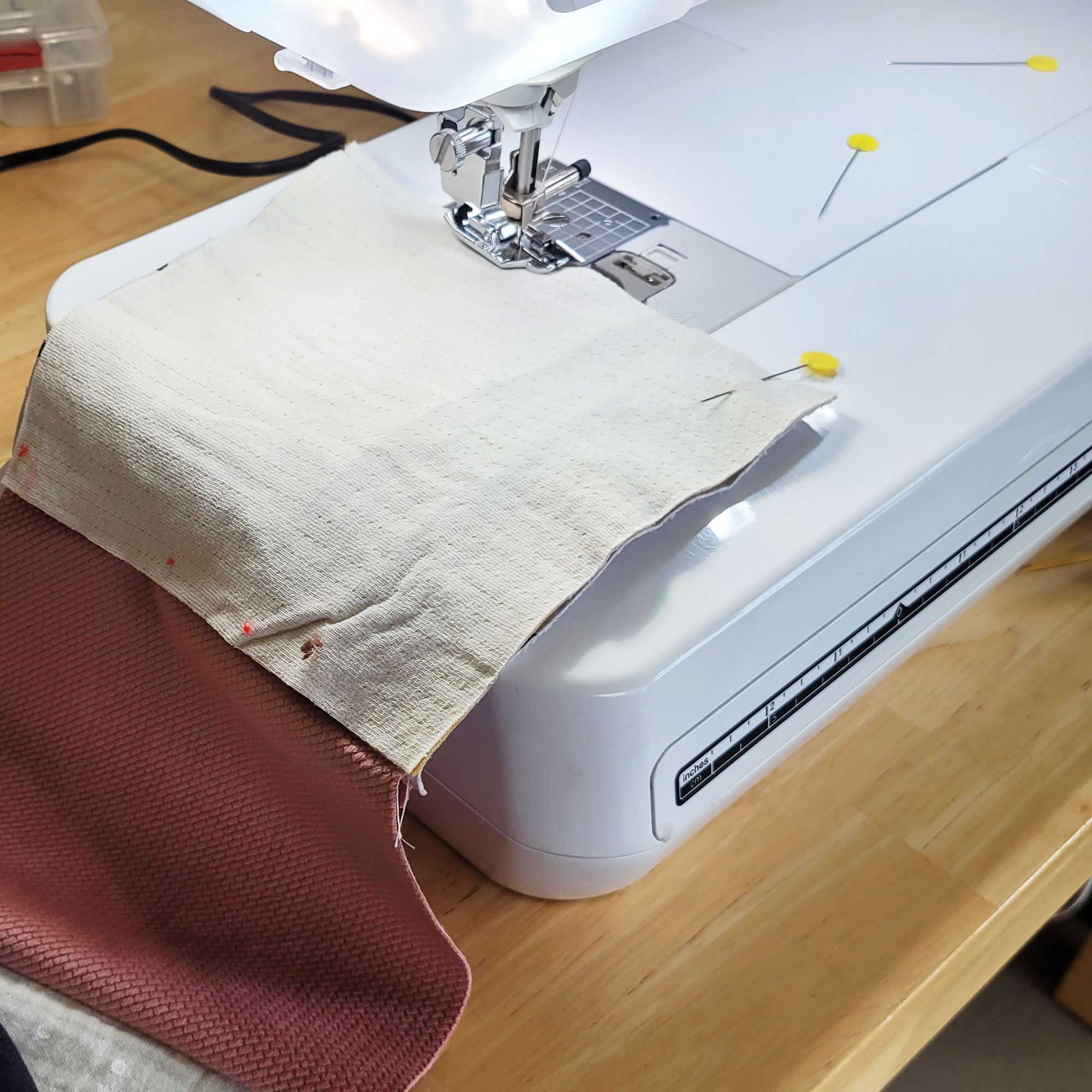 Weitere Quadrate sind zum Zusammennähen zusammengesteckt und unter die Nähmaschine gelegt. Das obere Quadrat ist diesmal grau und mit einer langen Stecknadel mit gelbem, flachen Kopf zusammengesteckt. Das Quadrat ist zu etwa 1/3 zusammengenäht. Eine weitere Stecknadel mit gelbem Kopf liegt rechts auf der Arbeitsfläche der Nähmaschine.