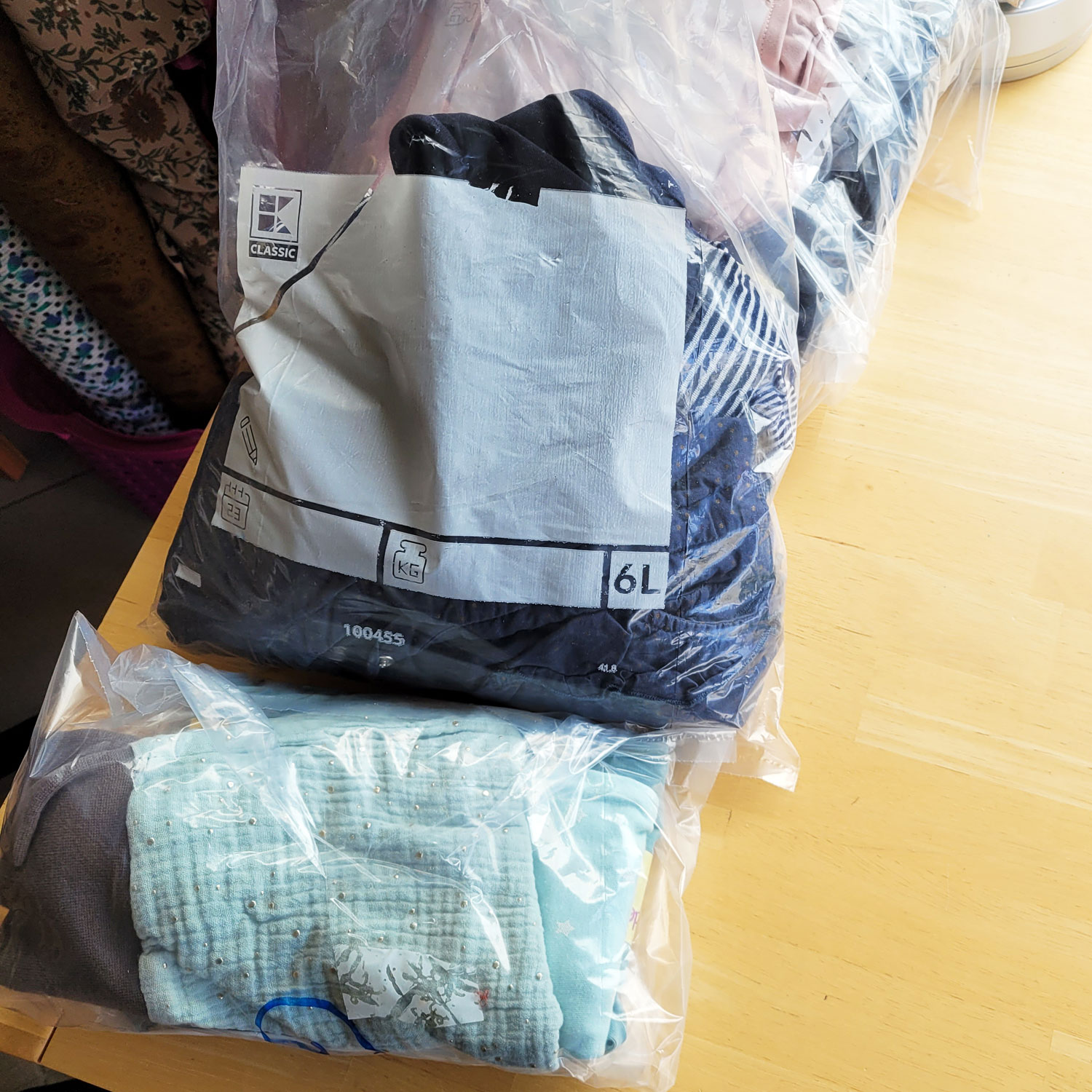 In 6l großen Gefriertüten befinden sich nach Farben sortiert Babysachen. Auf dem Bild zu sehen sind drei Tüten, die von unten nach oben folgende Farben enthalten: Grün, Blau, Rosa. Sie liegen auf einem hellbraunen Schreibtisch. In der linken oberen Bildecke sieht man einige Stoffrollen in Braun- und Beigetönen.