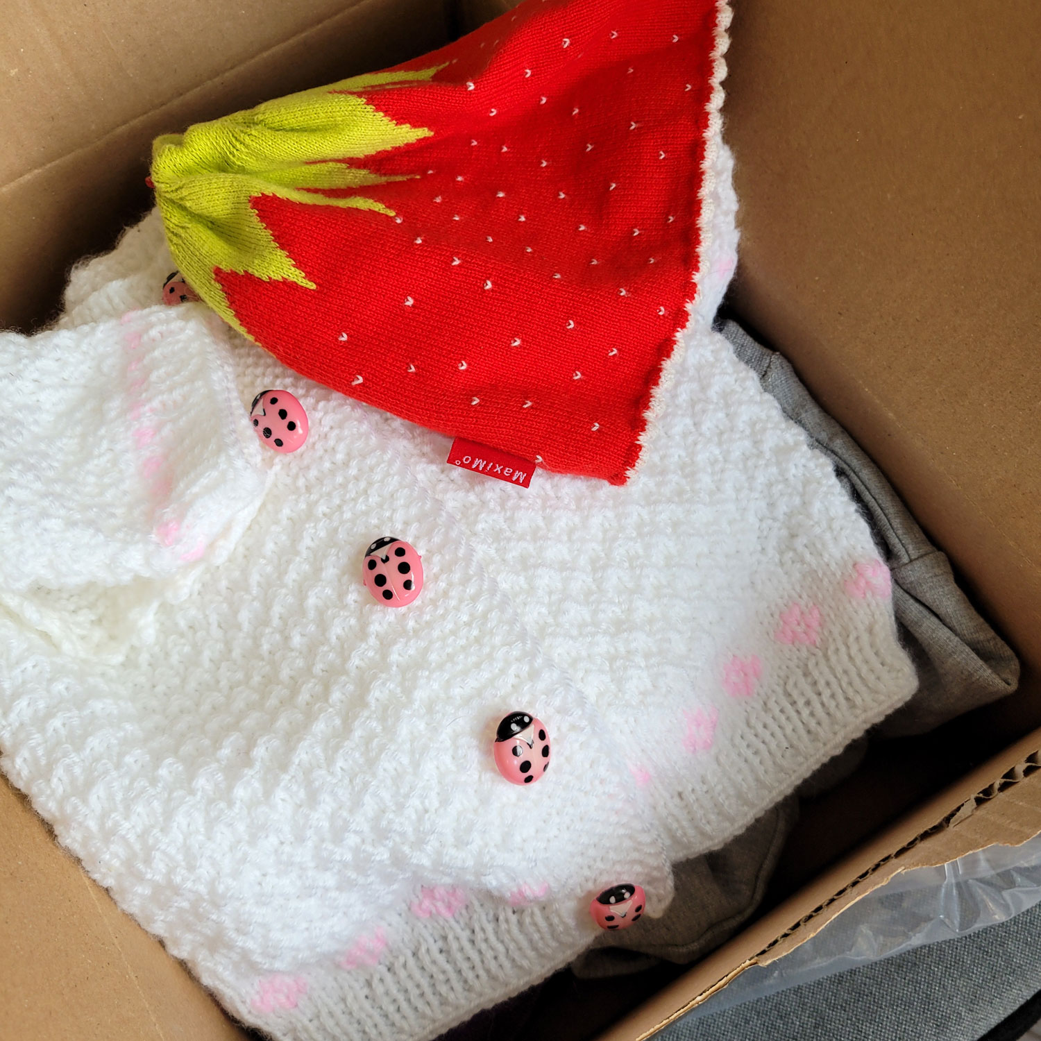 In einem Karton liegen eine kleine Babymütze in grün und rot, die aussieht wie eine niedliche Erdbeere und ein weißes Strickjäckchen mit rosa Marienkäferknöpfen