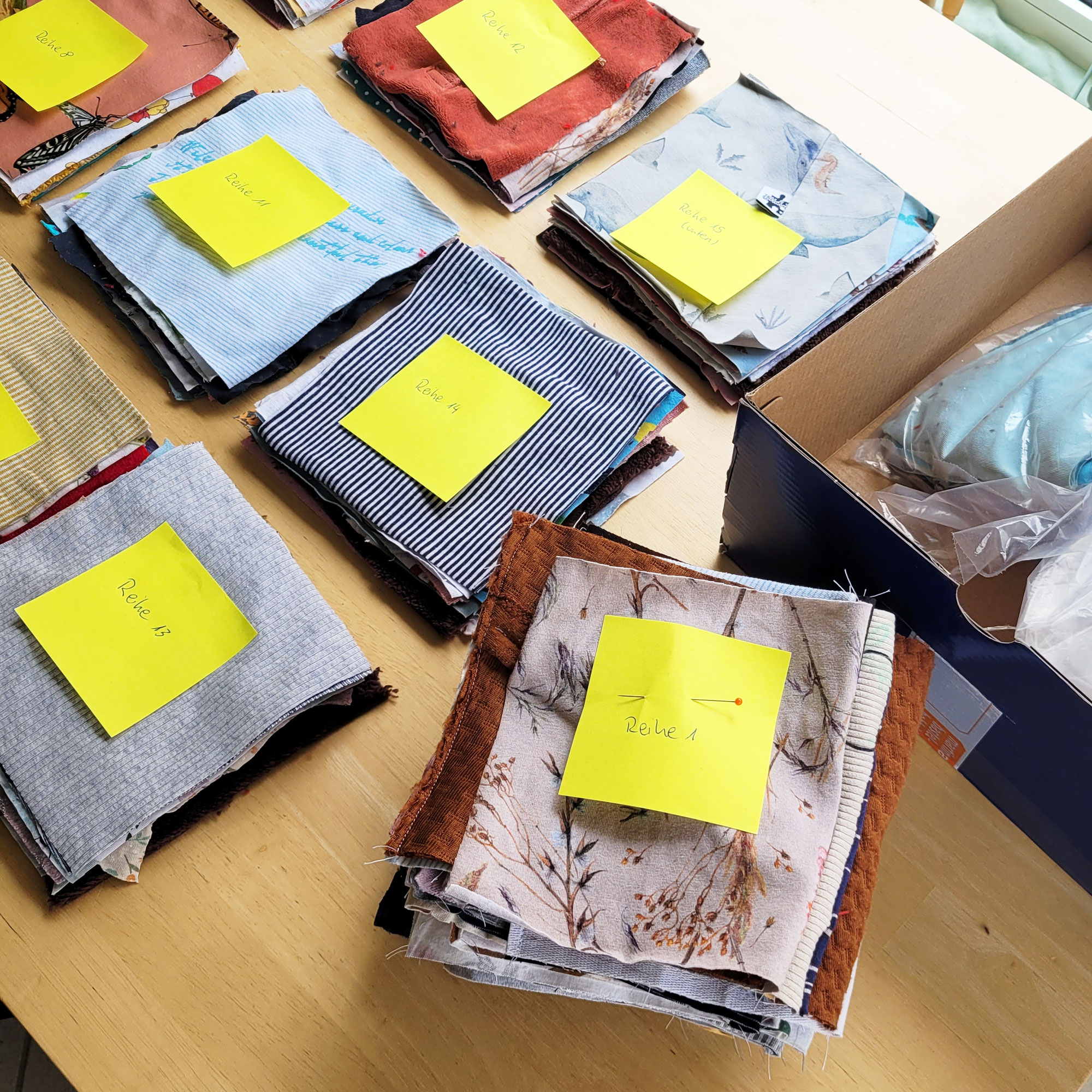 Auf einem hellen Schreibtisch aus Holz liegen 9 Häufchen aus Stoffquadraten. Auf jedem Päckchen klebt ein gelber Post-It Zettel mit einer Reihennumer. Der Haufen vorne Rechts ist höher, hier liegen mehrere Reihenhäufchen übereinander, der obere Haufen hat die Nummer Reihe 1. Die einzelnen Quadratstapel links sind Nummeriert von Reihe 8 bis 15. Es sind nicht alle Zahlen sichtbar.