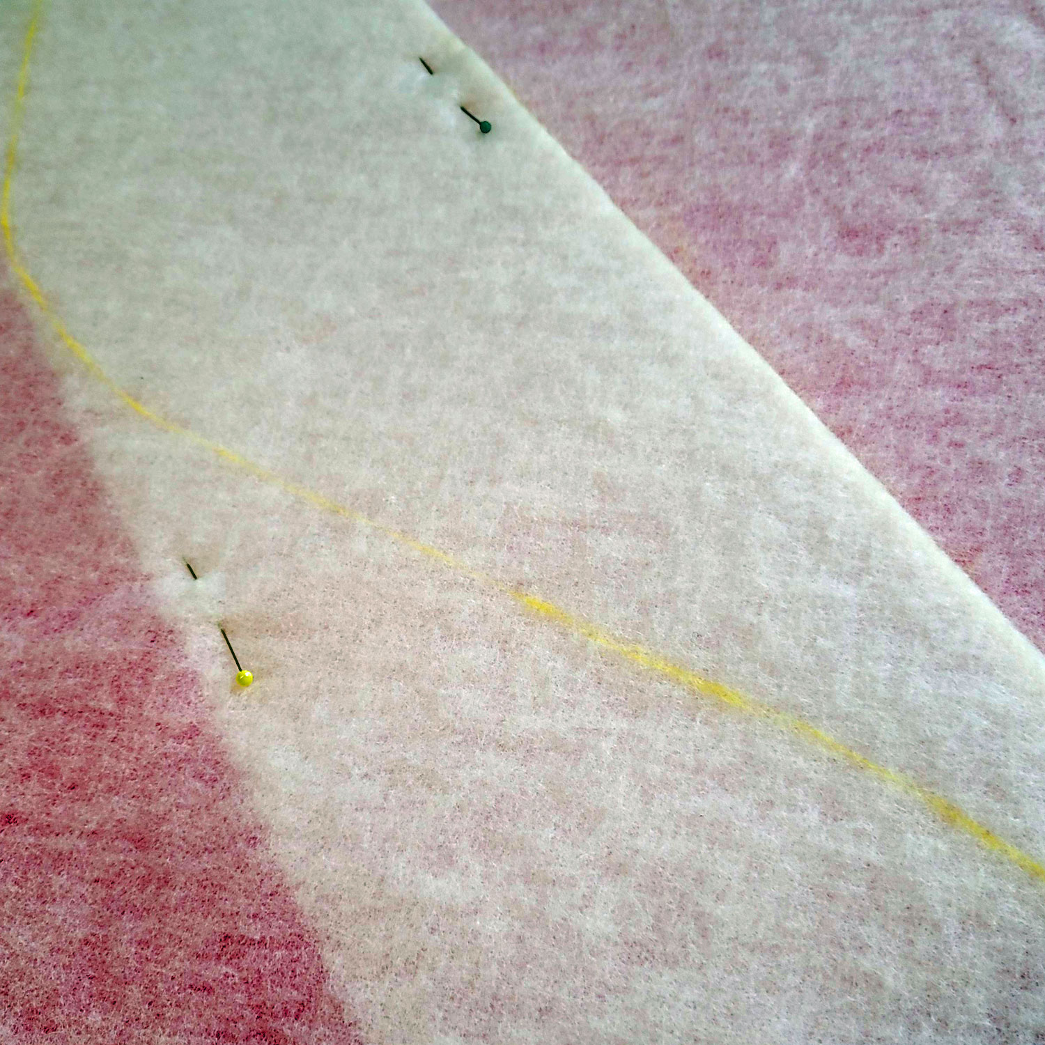 Zwei Lagen Vlieseline H630 sind mit Stecknadeln aufeinander gesteckt und überlappen sich um ca. 20 cm. Versetzt sieht man 2 Stecknadeln, die die Lagen halten. Durch die Vlieseline scheint pinkfarben die Schneidematte des Zuschneidetisches durch. Auf der überlappten Linie ist in gelber Kreide eine Wellenlinie eingezeichnet.