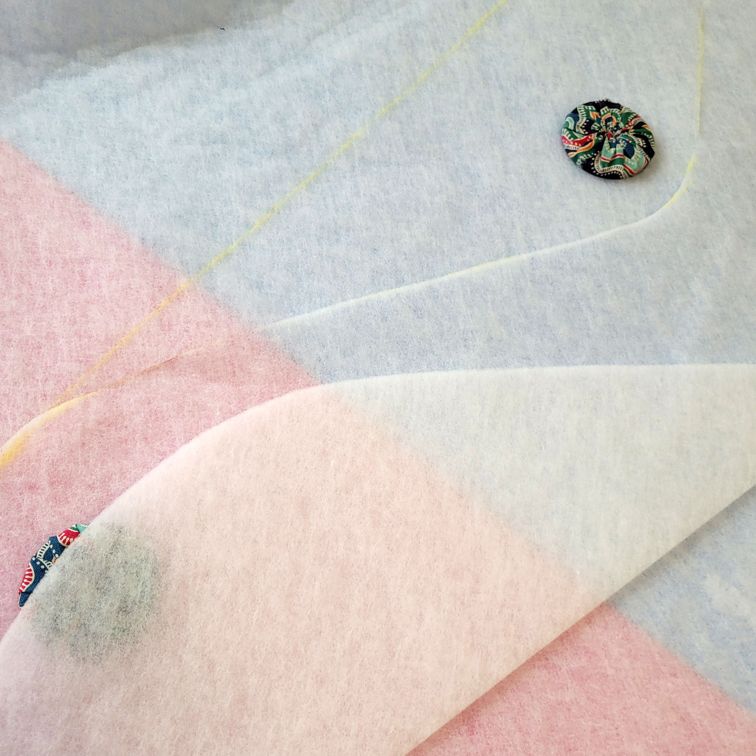 Die Vlieseline liegt auf dem Zuschneidetisch, man sieht die gelbe Wellenlinie teilweise . Auf der Vlieseline liegen zwei bunt mit Stoff bezogene Gewichte. Im Vordergrund liegt ein vom oberen Vlieselinestreifen abgeschnittenes Stück H630.