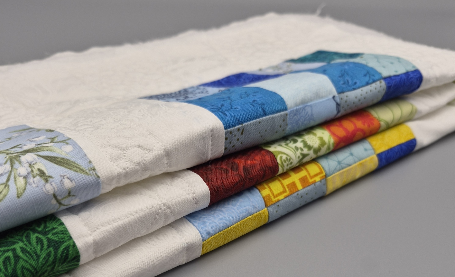 Auf einem Stapel vor grauem Hintergrund liegen drei Tischsets die in unterschiedlichen Stadien der Fertigstellung sind. Das untere ist in blau-gelb gehalten, das in der Mitte in grün-rot und das oben in blautönen